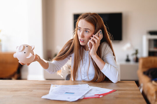 Woman talking on phone while holding piggy bank and looking concerned about financial issues