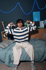 Teenage Black boy sitting on bed flexing arms and showing biceps, looking confidently at camera in modern bedroom with neon lights and coding themed decorations visible