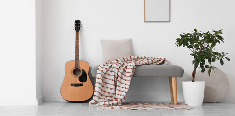 Soft bench with plaid, houseplant and guitar near white wall in room