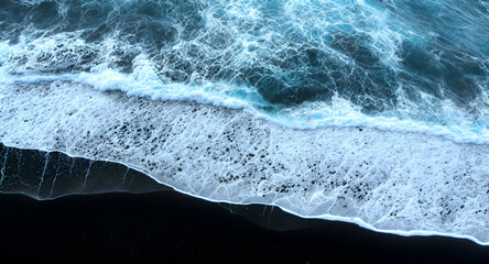 A dramatic aerial view of powerful ocean waves crashing onto a deep black-sand shoreline.