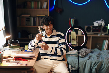 Portrait of young Black man smiling and holding trophy while sitting at desk in bedroom, pointing at award with finger, recording video using smartphone and ring light for social media