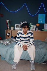Portrait of teenage Black boy sitting on bed with hands clasped, looking into camera, modern...