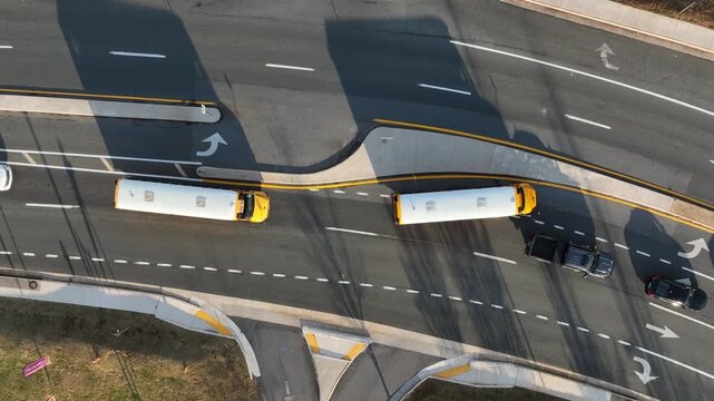 Top-down aerial view of two yellow school buses driving along multilane American roadway on a sunny day, with clear lane markings, turning arrows, moving cars. Peaceful afternoon in fall season.