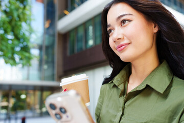 Young, beautiful happy Asian woman in green shirt look up, smile with confidence, holding smartphone and coffee cup in front of modern office building. City lifestyle, self care and mindfulness