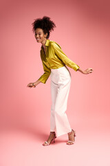 Bright and joyful young woman dancing in a chic outfit against a pink background