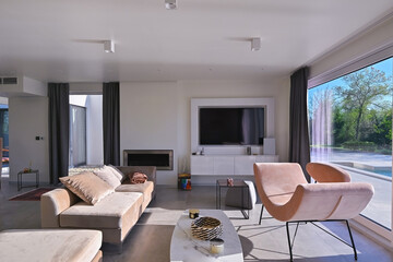 Obraz premium Modern luxury living room interior at sunset. Open-plan space featuring beige sofas, marble dining table, large glass windows with sun flare, and view of a patio. Stylish contemporary home design