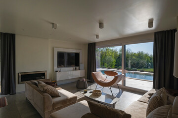 Obraz premium Modern luxury living room interior at sunset. Open-plan space featuring beige sofas, marble dining table, large glass windows with sun flare, and view of a patio. Stylish contemporary home design