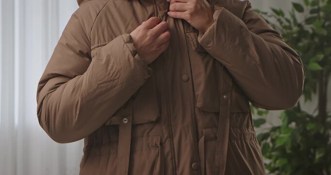 A woman buttons up her brown insulated winter jacket indoors, close up of her shortened stomach