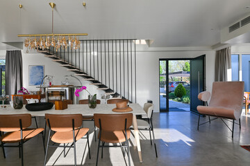 Modern luxury open-plan interior featuring a floating staircase, marble dining table, wooden kitchen, and beige sofa. Spacious contemporary home design with elegant lighting and gray tiled floors