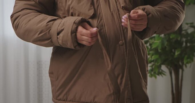 A woman ties the belt on her brown winter down jacket, close up of her stomach, preparing for cold weather indoors.
