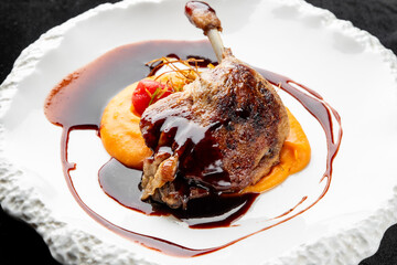 Crispy duck confit leg served on a bed of vibrant orange sweet potato or pumpkin puree, drizzled with a rich dark sauce on a modern white plate