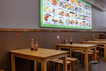 Interior of small Chinese cafe in Sanya, Hainan, with simple wooden tables and stools beneath large illuminated menu board showing photos of dishes. Quiet, everyday local eatery atmosphere.