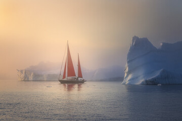 View of sailboat with red sails navigates through icy waters besides colossal icebergs under a hazy, golden sky, Greenland.