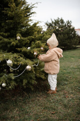 Cute funny baby girl 1-2 year old decorating Christmas tree outdoors. Winter holiday season. Childhood. Kid wearing fluffy teddy coat