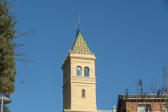View of a tall, ornate, beige tower with a green and white patterned roof reaches towards a clear blue sky, a blend of architectural styles, Barcelona, Catalonia, Spain.