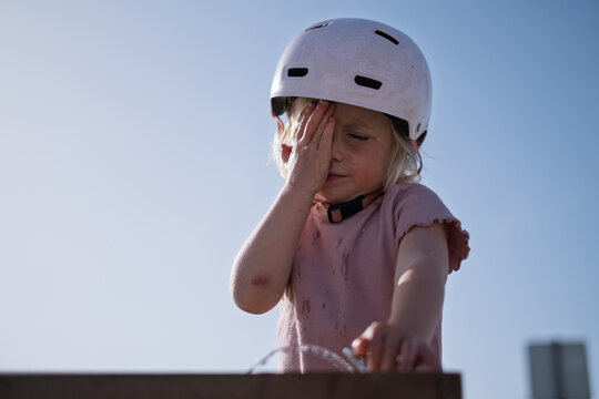 A little girl wearing a helmet drinks water from a public fountain and wipes her face. 