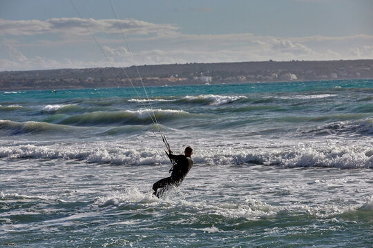 Man kiteboarding in choppy turquoise waters near Palma de Mallorca, riding strong wind and waves in full motion under a cloudy sky. thrill