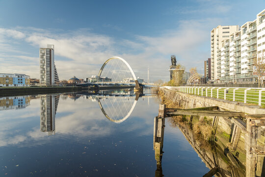 View of a serene river reflecting the modern architecture and iconic bridge under a vast blue sky, creating a striking contrast of urban textures, Glasgow, Scotland, United Kingdom.