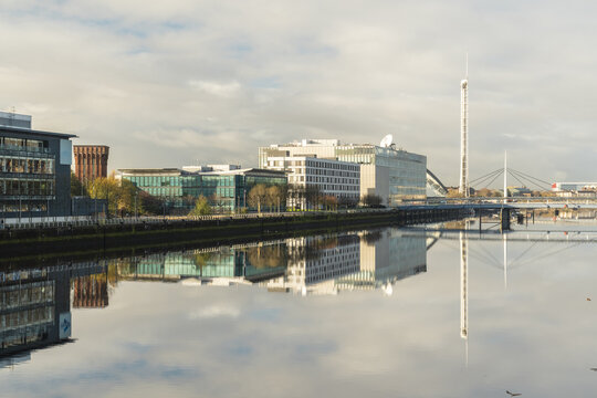 View of tranquil waters reflecting modern buildings and a towering structure under a soft, diffused sky, casting a serene light, Glasgow, Scotland, United Kingdom.