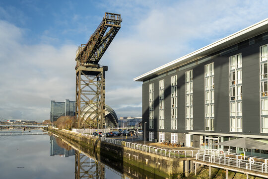 View of the Finnieston Crane stands tall against the blue sky and calm waters, contrasting with the modern building's clean lines, Glasgow, Scotland, United Kingdom.