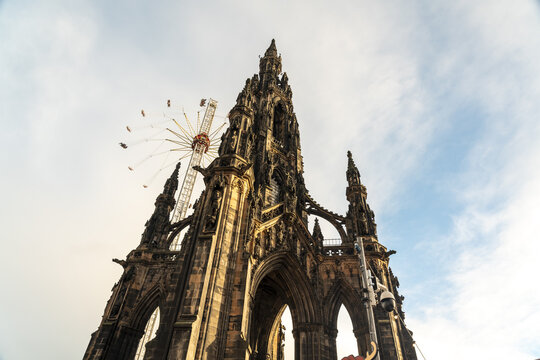 View of the Gothic spire of the Scott Monument rises dramatically against a backdrop of swirling clouds and a distant fairground ride, Edinburgh, Scotland, United Kingdom.