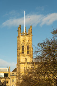 View of a golden sandstone tower of St John's Episcopal Church with Scottish flag fluttering atop, framed by the stark branches of winter trees, Edinburgh, Scotland, United Kingdom.