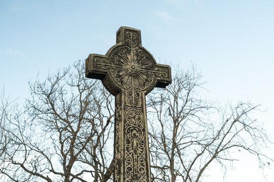 View of a weathered Celtic cross monument etched with intricate patterns stands tall against the pale blue sky, framed by bare tree branches, Edinburgh, Scotland, United Kingdom.