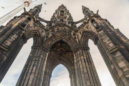 View of the Scott Monument's dark, gothic arches reach skyward, juxtaposed against the pale sky and a distant, spinning fairground ride, Edinburgh, Scotland, United Kingdom.