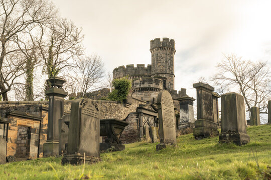 View of aged gravestones stand amidst the Old Calton Cemetery's green slopes, crowned by a stone tower under a muted sky, Edinburgh, Scotland, United Kingdom.