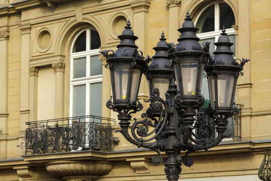 View of ornate black metal lamppost standing out against the pale yellow building facade with arched windows and wrought iron balcony, Frankfurt am Main, Hessen, Germany.