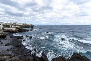 View of the rugged, dark volcanic rocks contrasting with the frothy white waves crashing against the shore near the white buildings, Tenerife, Canary Islands, Spain.