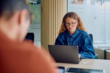 Woman with eyeglasses focused on her laptop, busy working at a desk in a contemporary co-working office space