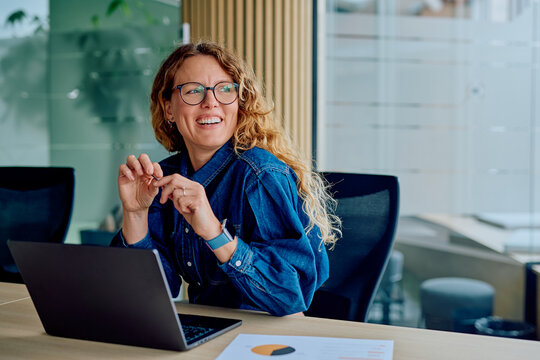 Smiling businesswoman laughing during an office conversation, sitting at a desk with a laptop and documents, expressing joy and confidence