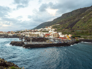 View of the town's colorful buildings nestled against the dark, rugged cliffs overlooking the turquoise sea, contrasting textures and hues creating a captivating scene, Garachico, Tenerife, Spain.