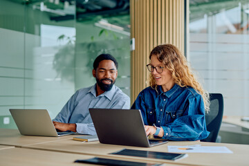 Two diverse colleagues smiling and working together on laptops at a modern office desk, sharing ideas and achieving business goals © we.bond.creations