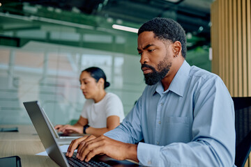 Serious male employee working diligently at laptop in a professional open-plan office setting, focusing on work with colleague