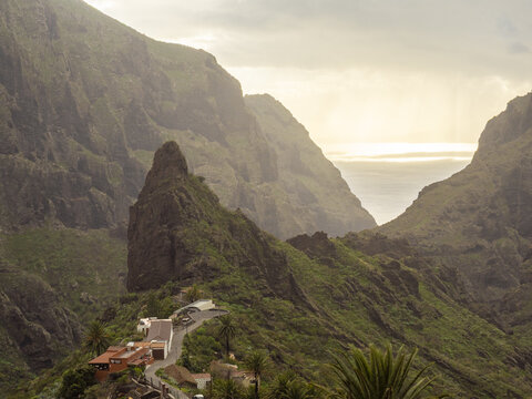 View of jagged cliffs rise dramatically, casting long shadows over the winding road and quaint village nestled below, a landscape of raw beauty, Masca, Tenerife, Spain.