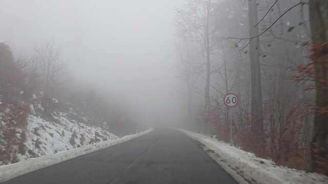 View from a car driving along a winding mountain road in late autumn with bare trees, light fog, and patches of snow on the roadside.