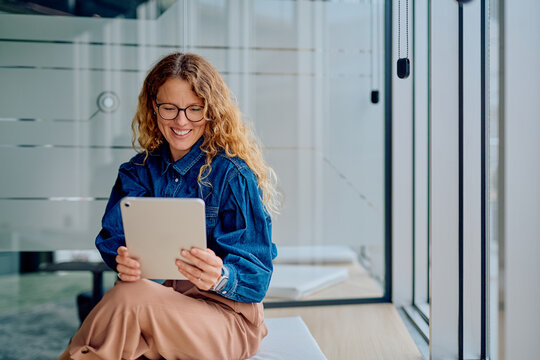 Woman smiling and engaging with a digital tablet, sitting casually in a modern office environment, connecting with technology