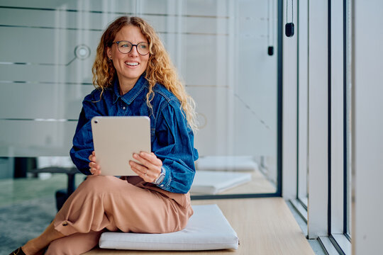 Smiling woman with curly hair and glasses using a tablet, working remotely from a modern relaxed office environment