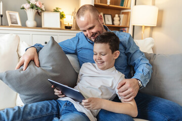 Shot of a handsome young man sitting on the sofa and bonding with his son while using a tablet. Father with teenage son browsing social media content using digital tablet in living room.