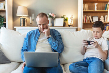 Shot of a cheerful little boy and his father using a smartphone and a digital tablet while being seated on a sofa at home during the day.