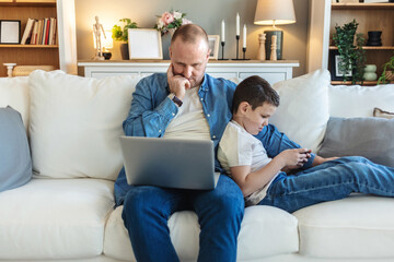 Shot of a cheerful little boy and his father using a smartphone and a digital tablet while being seated on a sofa at home during the day.