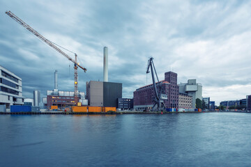 View of the Stadthafen's industrial architecture reflecting in the water, cranes reaching towards the sky under a cloudy twilight, Muenster, Germany.