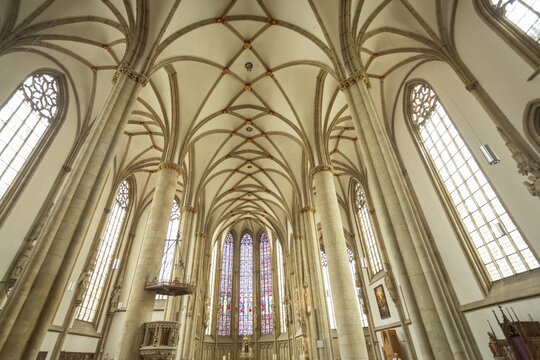 View of rows of wooden pews leading to a stained-glass window in Lambertikirche church with high vaulted ceilings and stone columns, Muenster, North Rhine-Westphalia, Germany.