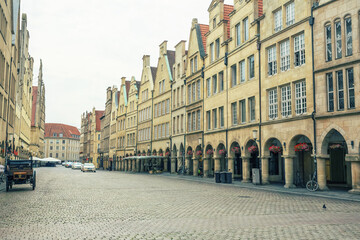 View of the street lined with tall, tan buildings with sharply angled roofs and arched entryways, a horse-drawn carriage on the cobblestone road, Muenster, North Rhine-Westphalia, Germany.