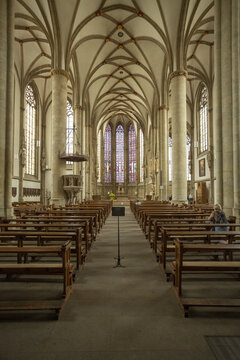 View of rows of wooden pews leading to a stained-glass window in Lambertikirche church with high vaulted ceilings and stone columns, Muenster, North Rhine-Westphalia, Germany.