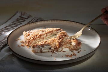 A slice of Napoleon cake rests on a plate, lit dramatically. A hand enters with a fork, ready to enjoy this decadent pastry in the soft light