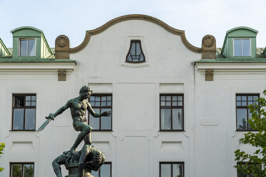 View of a bronze statue of a triumphant figure standing over a defeated foe, set against a backdrop of a white building with dark windows, Kalmar, Kalmar County, Sweden.