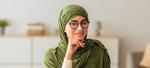 A young woman wearing a green hijab and glasses sits comfortably in a bright, minimalistic room. She rests her chin on her hand, presenting a thoughtful expression against a soft background.